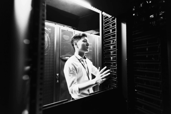 Black-and-white photo of a person in a white shirt and lanyard standing in a server room, interacting with a server rack. Multiple server cabinets are visible in the background, emphasizing a technical and professional environment. The image highlights the human role in maintaining IT infrastructure and managing data systems.