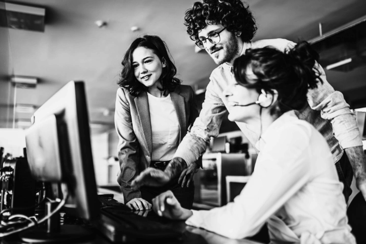 Three individuals in a modern office setting gathered around a computer monitor. One person is seated wearing a headset and pointing at the screen, while the other two stand nearby, attentively observing. The workspace includes desks, computers, and overhead lighting, capturing a moment of active collaboration and team communication.