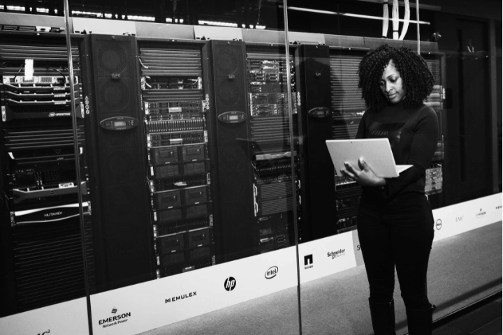 Person working on a laptop in front of a glass wall overlooking a secure server room filled with racks of enterprise-grade hardware. Technology logos from major vendors line the bottom of the glass, signaling trusted infrastructure partnerships. The image conveys a cybersecurity-focused environment, emphasizing vigilance, layered defense, and the human oversight behind resilient digital systems.