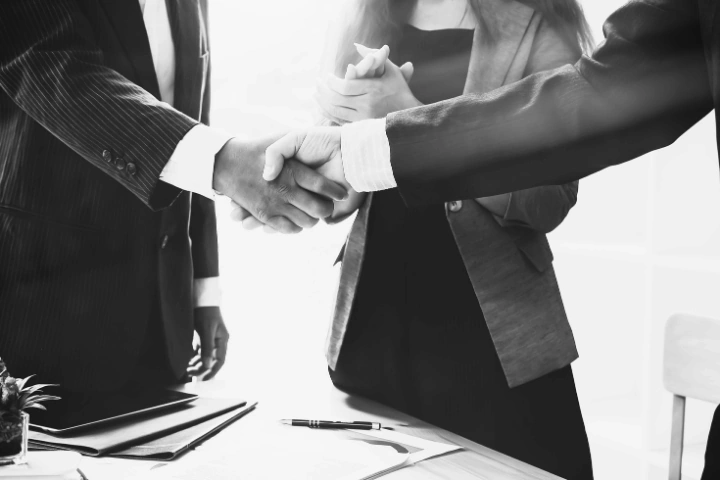 Black-and-white photo of three individuals in business attire engaged in a professional interaction. Two people are shaking hands across a table, symbolizing a formal agreement or partnership, while a third person stands nearby observing. Documents, a pen, and a small plant sit on the table in a modern office setting. The image conveys themes of trust, collaboration, and doing the right thing—emphasizing a commitment to one’s word and honoring professional relationships.