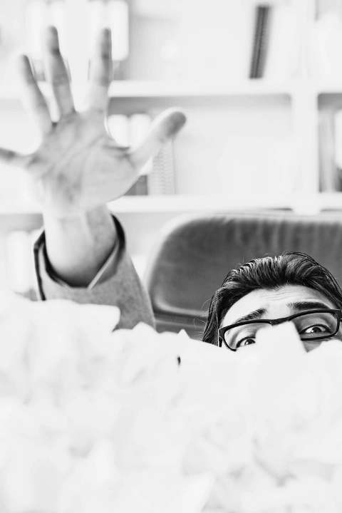Black-and-white image of a person partially buried under a pile of crumpled papers, with only their eyes and one raised hand visible. The person wears glasses and appears to be reaching upward, suggesting a sense of overwhelm or a plea for help. The blurred background includes a bookshelf and a chair, evoking an office or study setting and symbolizing stress or overload in a professional or academic environment.