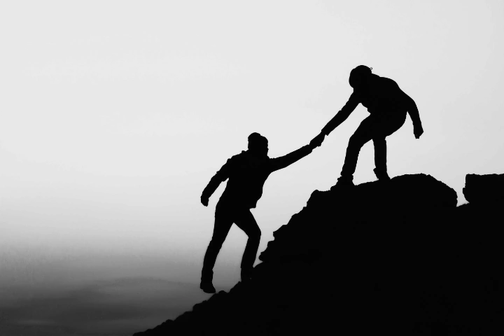 Black-and-white silhouette of two individuals on a steep rocky incline. One person stands higher, reaching down to help the other climb, symbolizing support, teamwork, and shared effort. The dramatic contrast and rugged terrain emphasize the importance of lifting others up and navigating challenges together with intention and care.