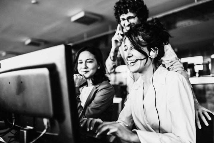 Three individuals in a collaborative office setting gathered around a computer monitor. One person is seated, wearing a headset and actively typing, while the other two stand nearby, focused on the screen. Their body language suggests engaged teamwork, possibly in customer support, tech troubleshooting, or a software development discussion. The environment includes desks, computers, and overhead lighting, reflecting a modern, professional workspace.