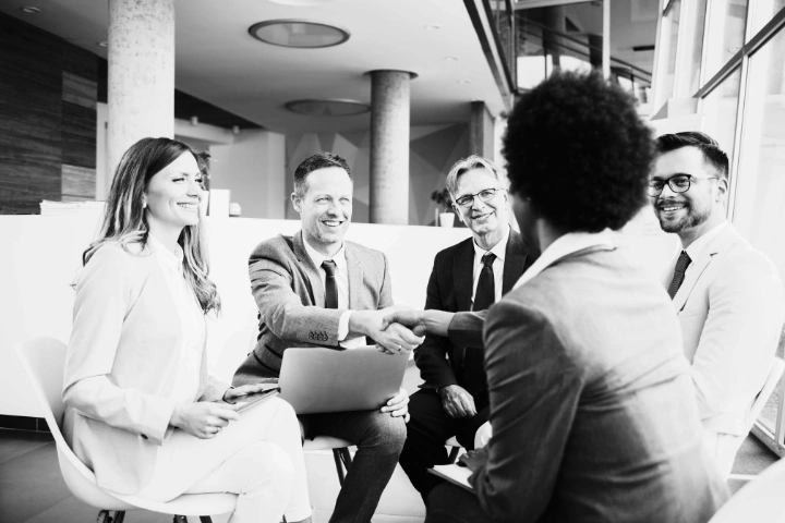 Group of five professionally dressed individuals seated in a modern office. Two people in the center are shaking hands, symbolizing a formal agreement or introduction, while others look on with engaged, smiling expressions. The setting features large windows, cylindrical columns, and contemporary furniture, conveying a collaborative and welcoming atmosphere. The image reflects themes of partnership, trust, and doing the right thing—emphasizing integrity, mutual respect, and commitment to one’s word in professional relationships.