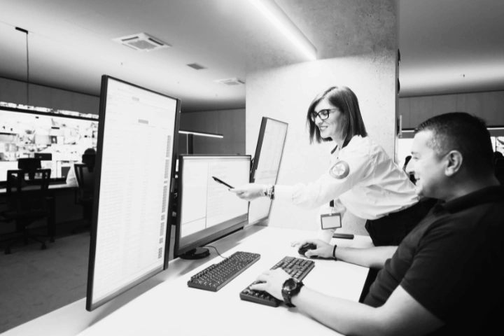 Modern security operations center (SOC) with two individuals working at a desk surrounded by multiple monitors. One person, wearing a uniform with a badge and lanyard, points at a screen with a pen while discussing with a seated colleague. The background includes additional workstations and surveillance feeds. The image offers a behind-the-scenes view into a SOC, highlighting real-time collaboration, incident response, and the infrastructure that protects digital environments.