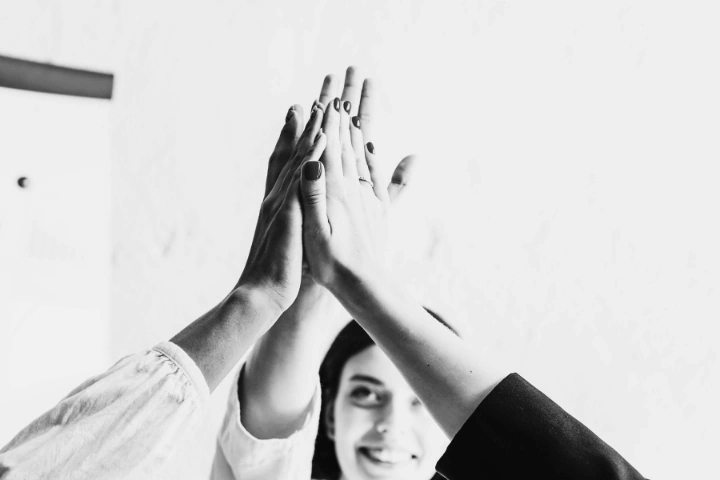 Black-and-white photo of four hands meeting in a high-five gesture, centered in the frame. One smiling face is partially visible in the background, set against a bright, minimalistic backdrop. The image conveys a sense of teamwork, celebration, and shared success.
