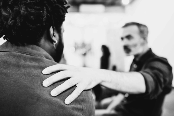Black-and-white photo of two individuals in conversation. The person in the foreground, wearing a dark shirt, has their back to the camera. The person in the background, slightly out of focus, places a hand on the other's shoulder in a gesture of support. The indoor setting suggests a moment of empathy, connection, or quiet encouragement.