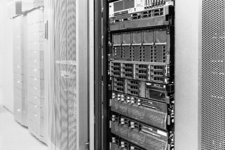 Close-up view of a server rack in a data center, showing multiple stacked servers with ventilation grills, indicator lights, and connection ports. Surrounding perforated metal panels support airflow and cooling. The image highlights the physical infrastructure behind digital services, cloud computing, and data storage.